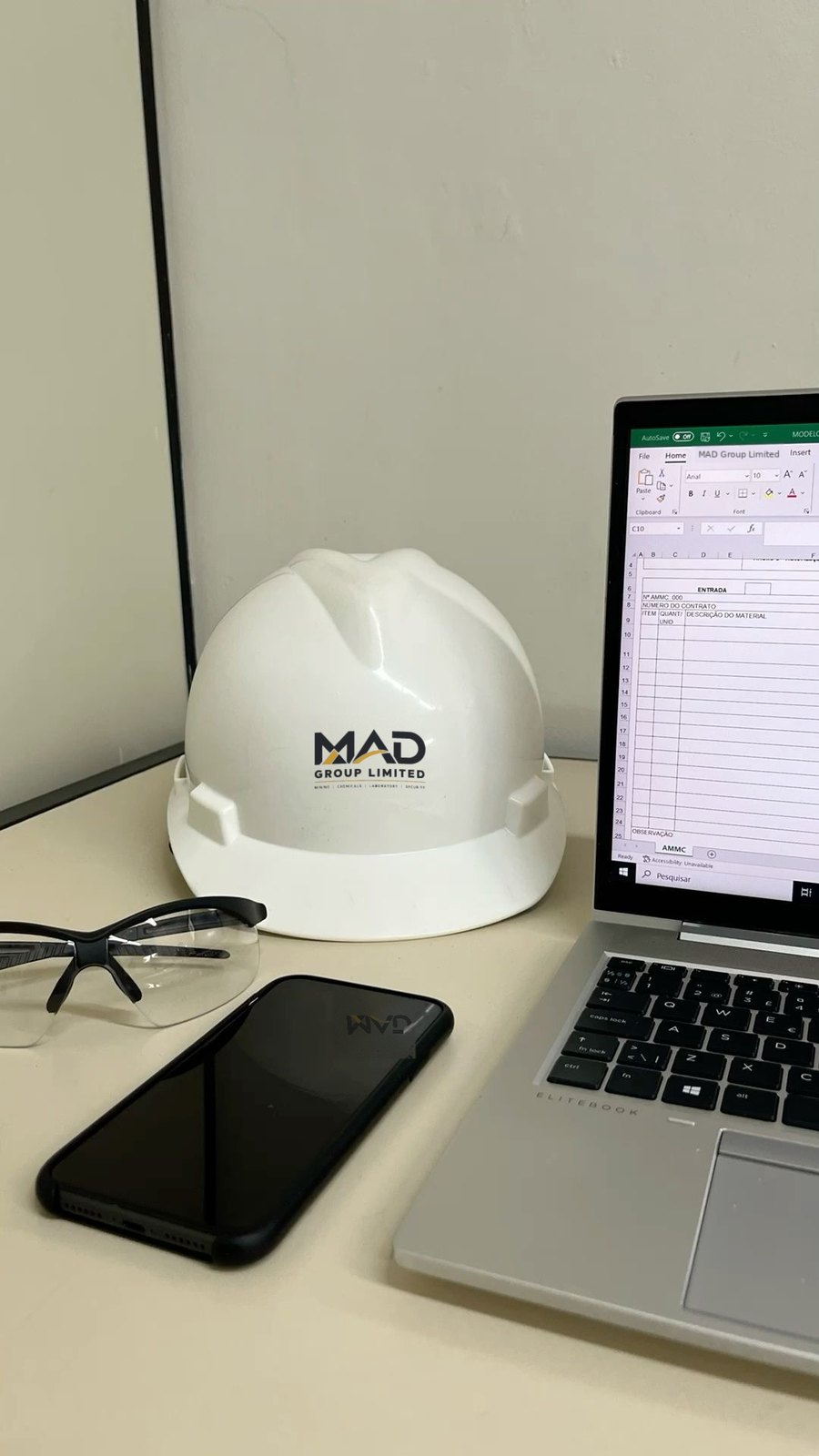 White safety helmet with the MAD Group Limited logo on a desk beside safety glasses, a phone and a laptop