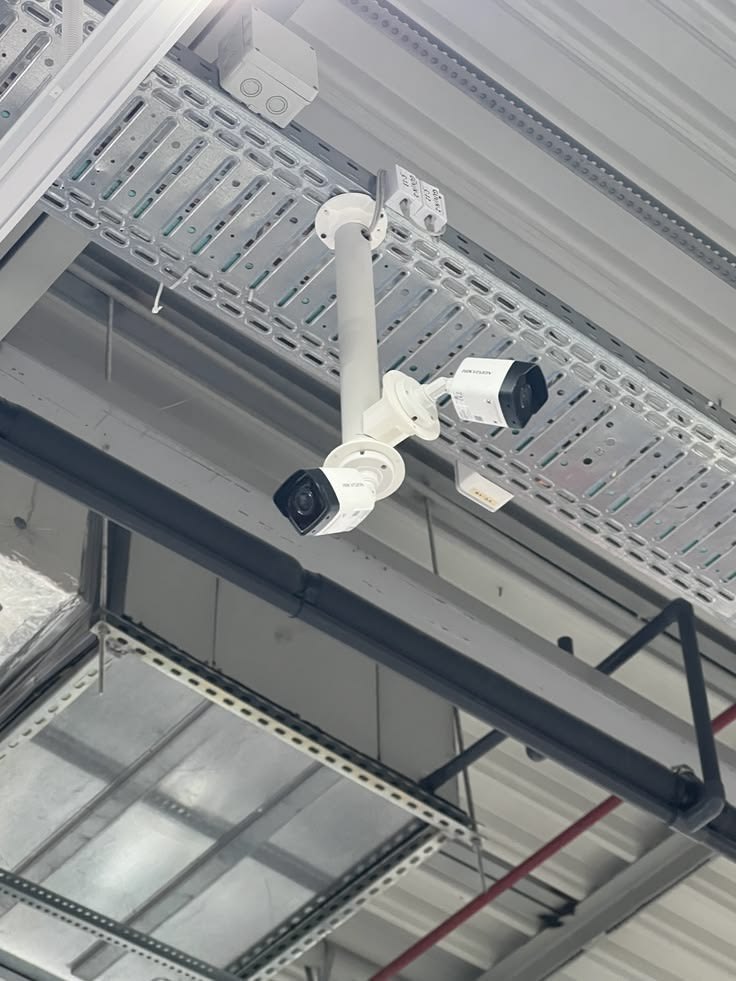Pair of CCTV cameras mounted on a ceiling bracket under cable trays inside a processing plant